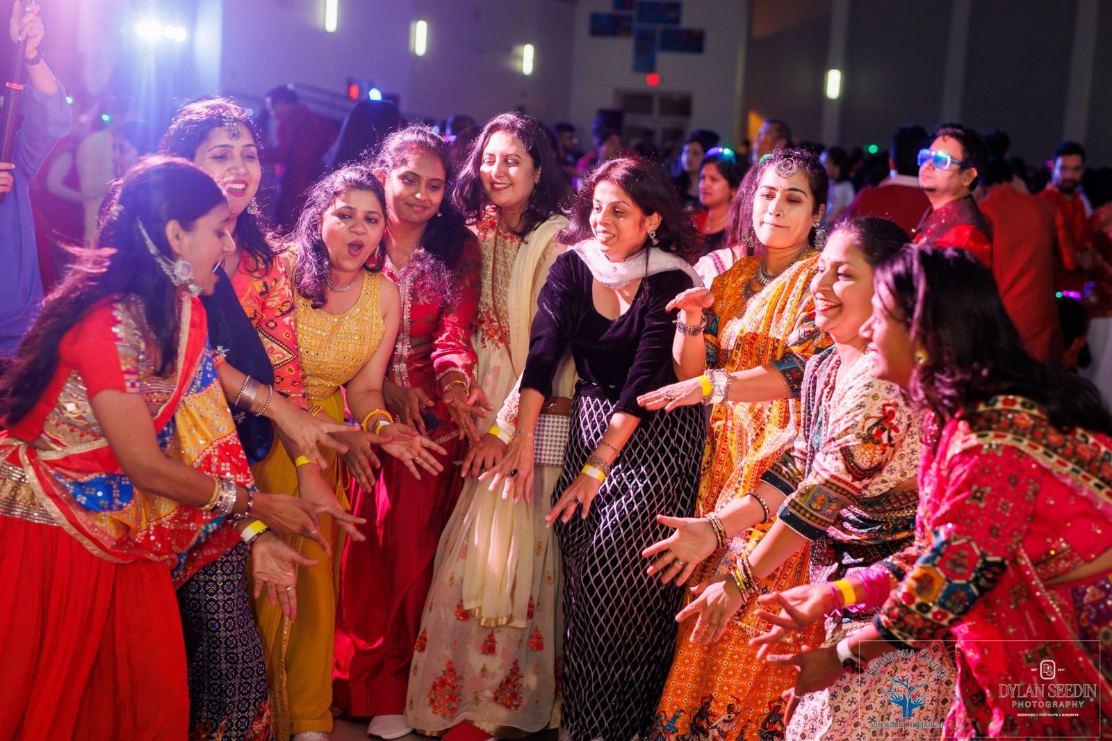 A circle of women in vibrant outfits dancing and clapping in a purple-lit hall