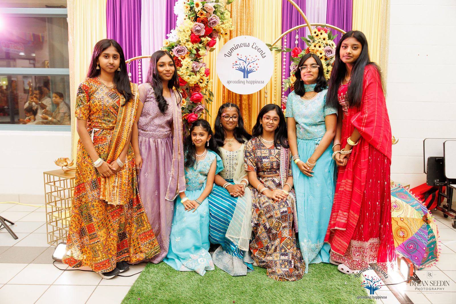 A group of girls and young teens in colourful traditional outfits at the floral backdrop